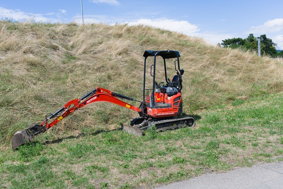 Mini ekskavatorius dirba ankštame kieme prie tvoros ir namo